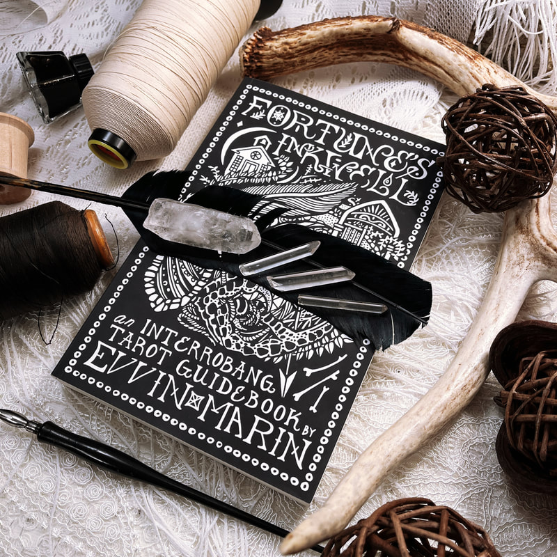 Fortune's Inkwell: an Interrobang Tarot Guidebook by Evvin Marin, photographed on a lace tablecloth with antlers, a quill feather, pen and ink supplies, spools of thread, and quartz crystals.
